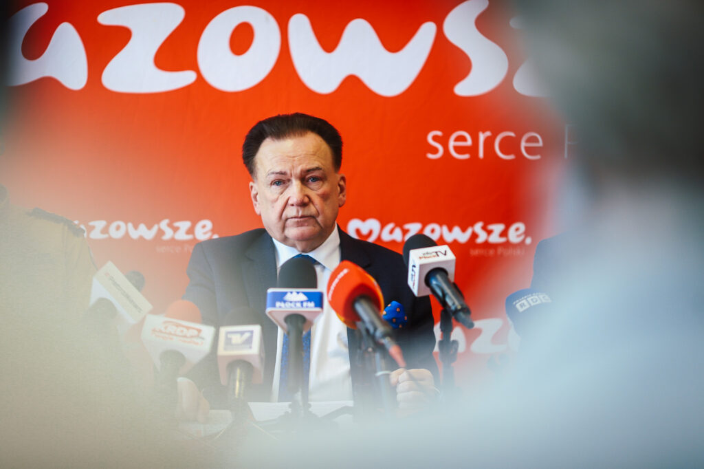 Polityk w garniturze wygłasza oświadczenie na konferencji prasowej dotyczącej zakończenia nielegalnego składowiska odpadów w Nowym Miszewie, otoczony mikrofonami na czerwonym tle z napisem Mazowsze.