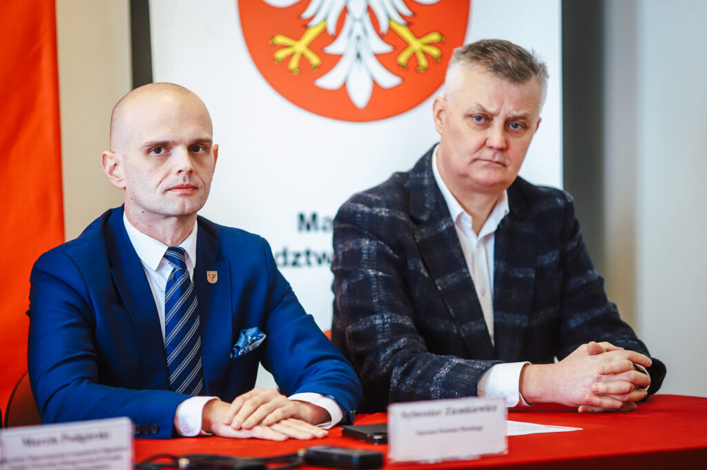 Dwóch mężczyzn w garniturach siedzi przy stole podczas konferencji prasowej w Nowym Miszewie, ogłaszającej koniec nielegalnego składowiska odpadów.