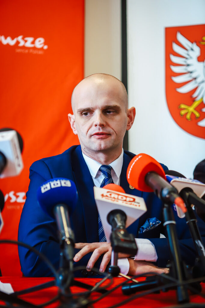 Mężczyzna w granatowym garniturze przemawia podczas konferencji prasowej, otoczony mikrofonami, na tle czerwonych banerów i godła, w kontekście zakończenia nielegalnego składowiska odpadów w Nowym Miszewie.