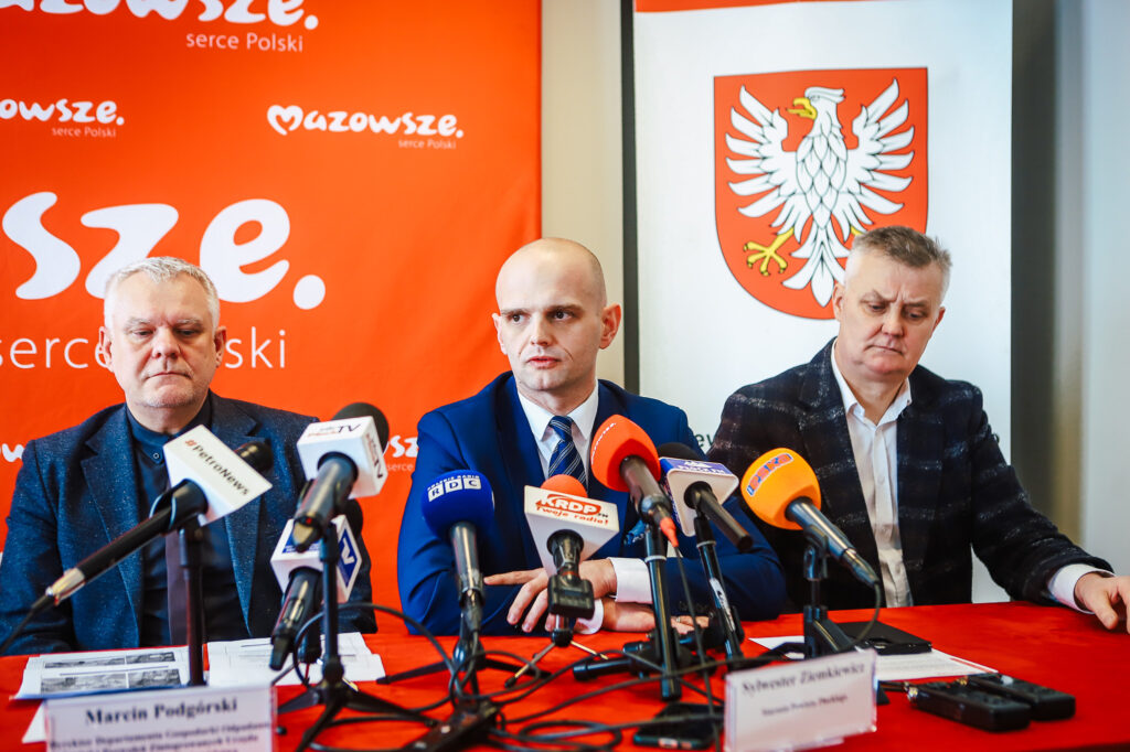 Trzej mężczyźni siedzą przy stole podczas konferencji prasowej w Nowym Miszewie, na tle czerwonego banera i herbu, informują o zakończeniu nielegalnego składowiska odpadów.