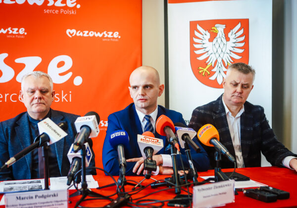 Trzej mężczyźni siedzą przy stole podczas konferencji prasowej w Nowym Miszewie, na tle czerwonego banera i herbu, informują o zakończeniu nielegalnego składowiska odpadów.