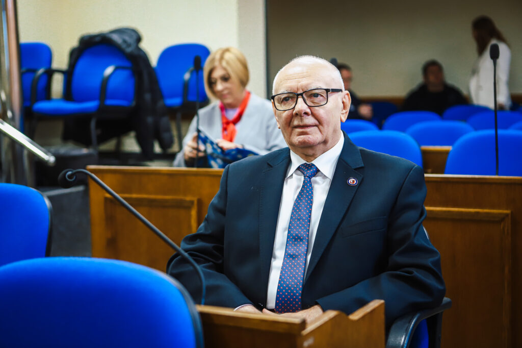 Uczestnik XIX sesji Rady Powiatu Płockiego w kadencji 2024–2029 siedzi przy pulpicie w sali sesyjnej, ubrany w garnitur i okulary, z mikrofonem przed sobą, a w tle kobieta czyta materiały.