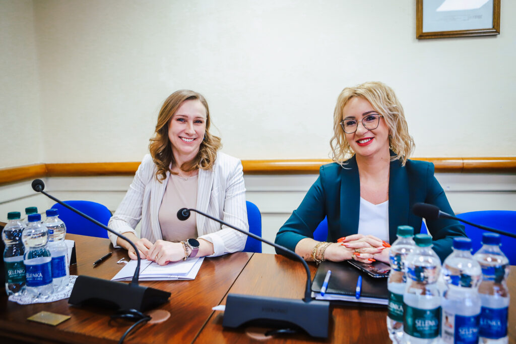 Dwie kobiety uśmiechnięte siedzą przy stole w sali konferencyjnej podczas XIX sesji Rady Powiatu Płockiego w kadencji 2024–2029, z mikrofonami, notatnikami i butelkami wody na stole.