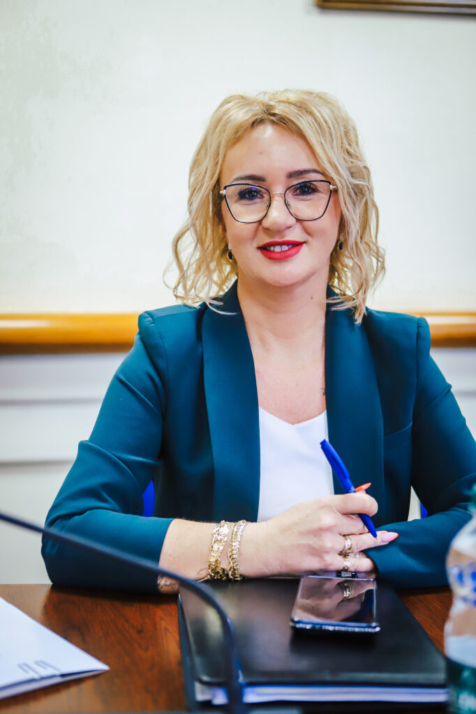 Uśmiechnięta kobieta z blond włosami i okularami w granatowym żakiecie siedzi przy stole podczas XIX sesji Rady Powiatu Płockiego w kadencji 2024–2029, trzymając niebieski długopis nad telefonem leżącym na notesie.