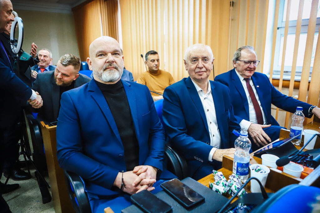 Trzech mężczyzn w niebieskich garniturach siedzi przy stole w sali sesyjnej podczas XIX sesji Rady Powiatu Płockiego w kadencji 2024–2029.