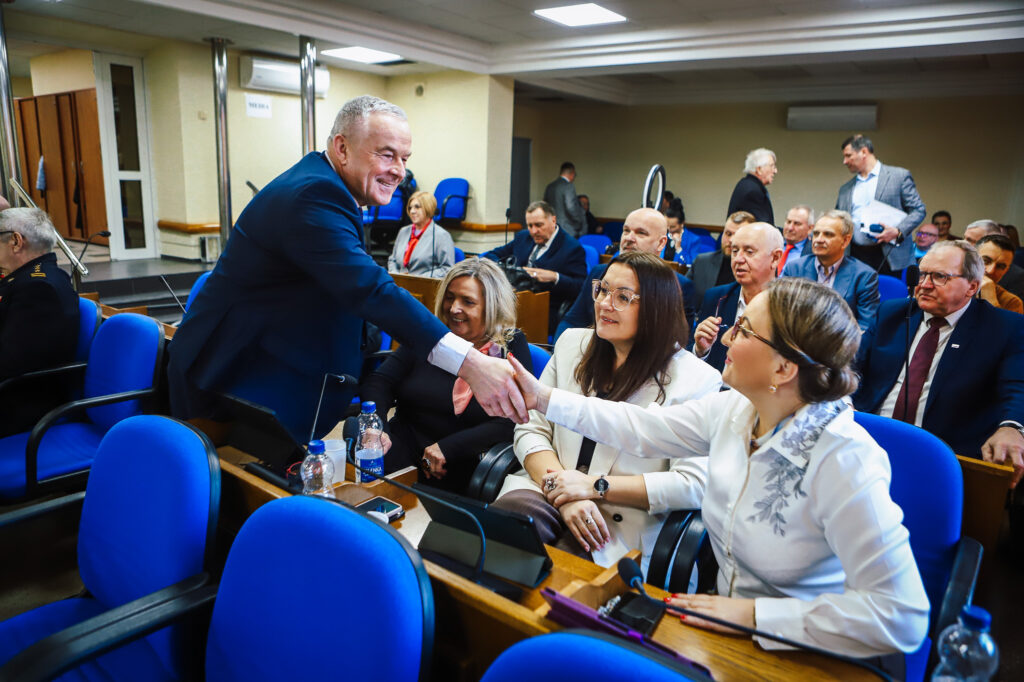 Na zdjęciu z XIX sesji Rady Powiatu Płockiego (kadencja 2024–2029) mężczyzna w granatowym garniturze podaje dłonie dwóm kobietom siedzącym na pierwszym rzędzie podczas obrad.