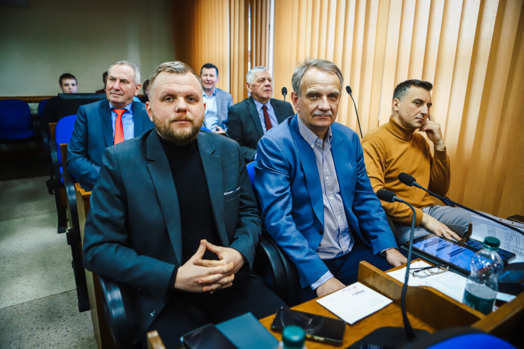 Grupa mężczyzn w sali obrad Powiatu Płockiego podczas XIX sesji Rady Powiatu w kadencji 2024–2029, siedzących przy biurkach z mikrofonami i dokumentami.