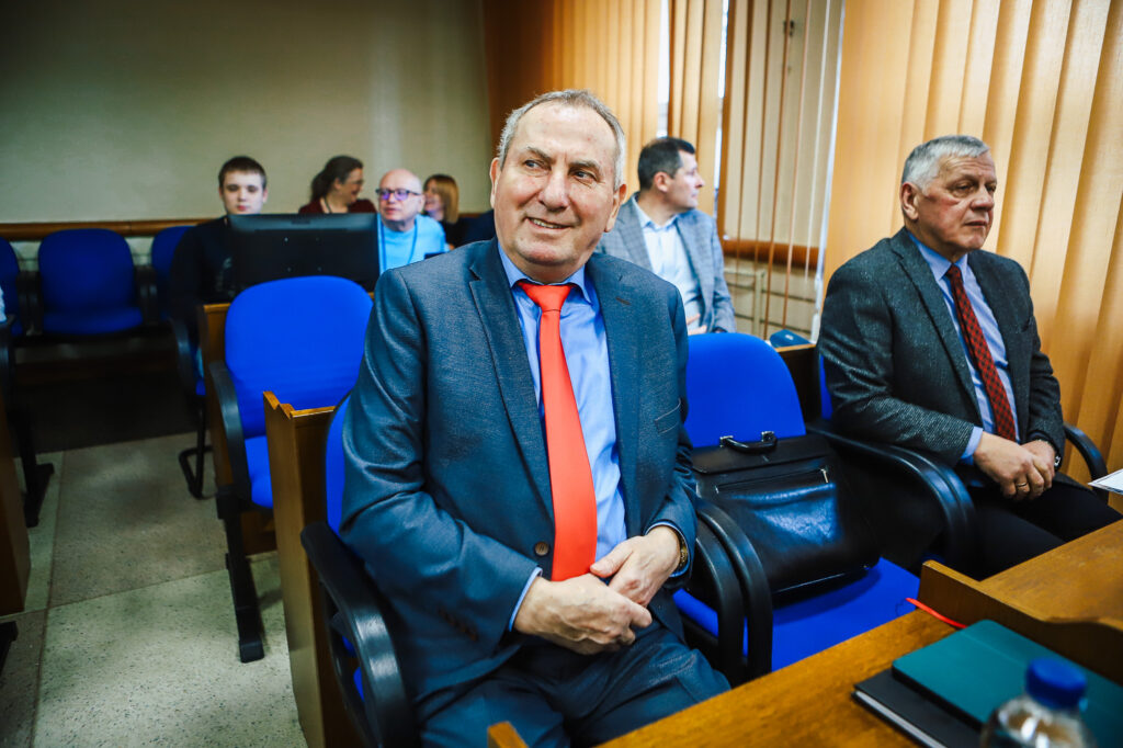 Uczestnicy XIX sesji Rady Powiatu Płockiego w sali obrad; na pierwszym planie mężczyzna w granatowym garniturze i czerwonym krawacie, uśmiechnięty.