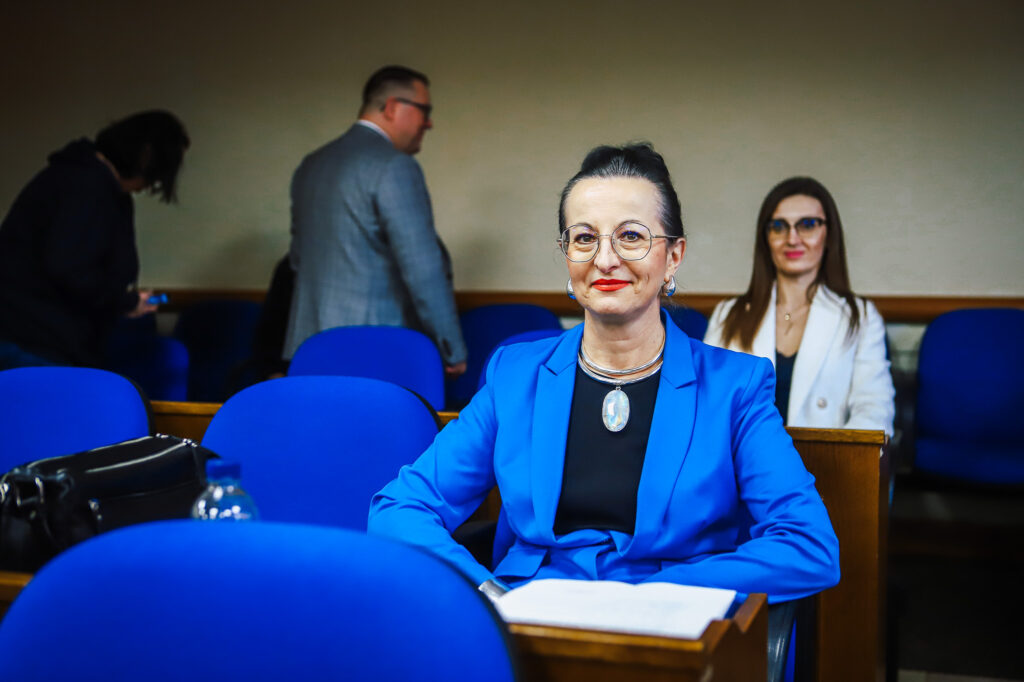 Uśmiechnięta kobieta w niebieskim garniturze siedzi na pierwszym planie w sali obrad podczas XIX sesji Rady Powiatu Płockiego w kadencji 2024–2029.