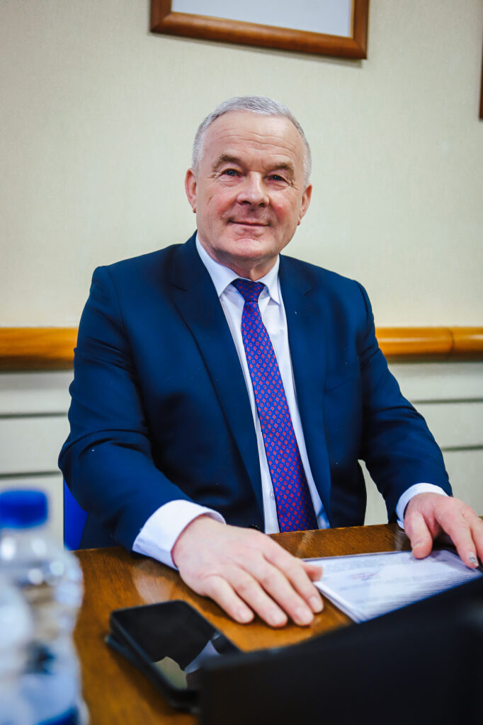 Mężczyzna w granatowym garniturze i czerwono-niebieskim krawacie siedzi przy stole podczas XIX sesji Rady Powiatu Płockiego w kadencji 2024–2029.