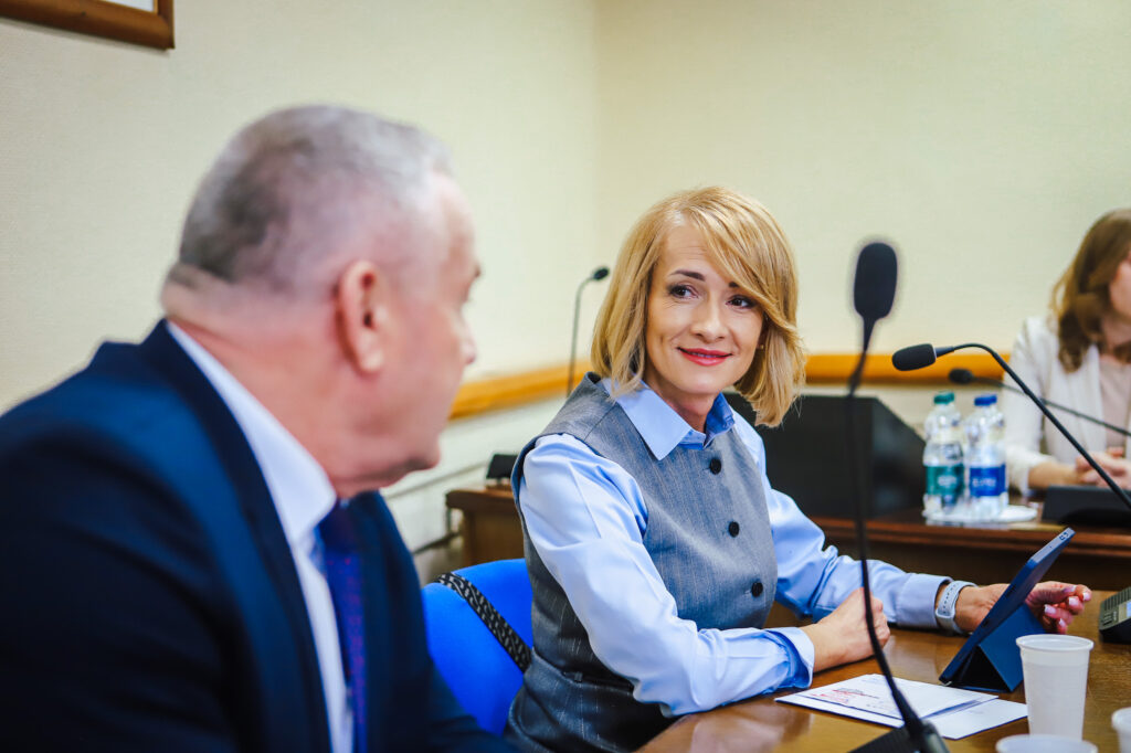 Kobieta z blond włosami w niebieskiej koszuli i szarym żakiecie siedzi przy stole z mikrofonem i tabletem podczas XIX sesji Rady Powiatu Płockiego (kadencja 2024–2029), uśmiechając się do rozmówcy.