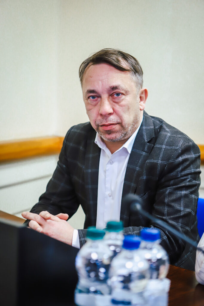 Mężczyzna w szarej kraciastej marynarce i białej koszuli siedzi przy stole z mikrofonem i butelkami wody podczas XIX sesji Rady Powiatu Płockiego w kadencji 2024–2029.