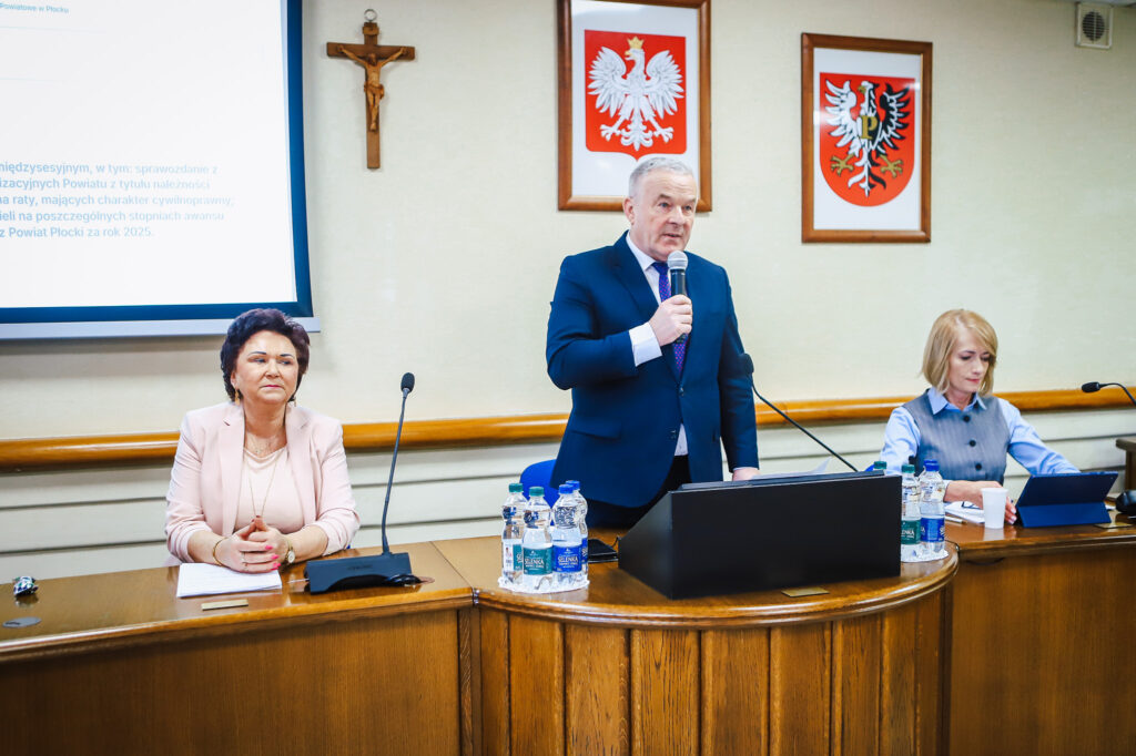 Podczas XIX sesji Rady Powiatu Płockiego w kadencji 2024-2029 mężczyzna w granatowym garniturze przemawia do mikrofonu stojąc przy mównicy, a po jego bokach siedzą dwie kobiety, w tle krzyż i herby na ścianie.