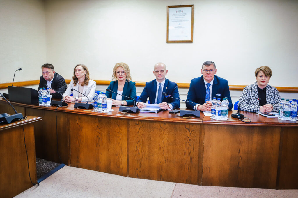 Sześciu radnych Rady Powiatu Płockiego w garniturach i marynarkach siedzi przy długim drewnianym stole w sali obrad podczas XIX sesji kadencji 2024–2029, z mikrofonami, dokumentami i butelkami wody.