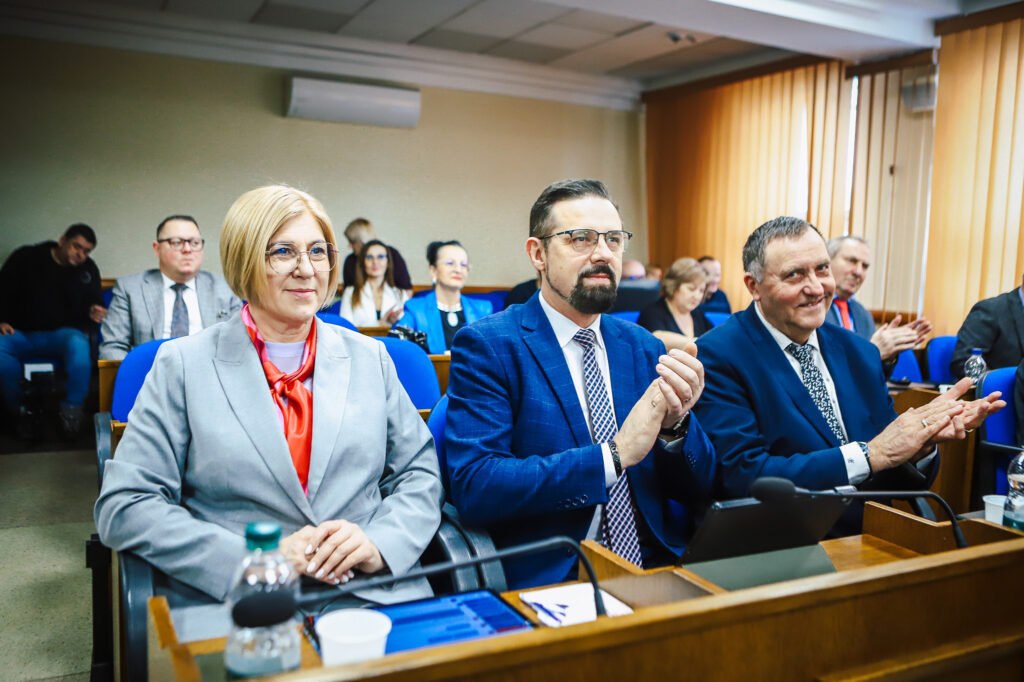 Trzy osoby w garniturach siedzą w pierwszym rzędzie podczas XIX sesji Rady Powiatu Płockiego w kadencji 2024–2029 i oklaskują.