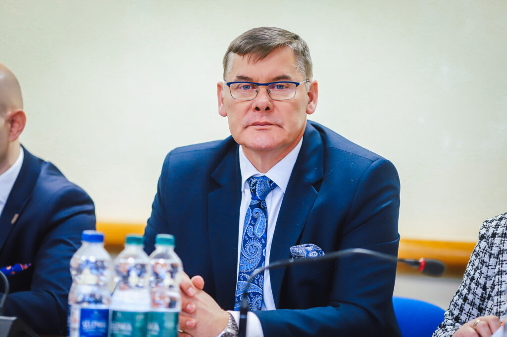 Mężczyzna w granatowym garniturze i okularach siedzi przy stole podczas XIX sesji Rady Powiatu Płockiego w kadencji 2024–2029.