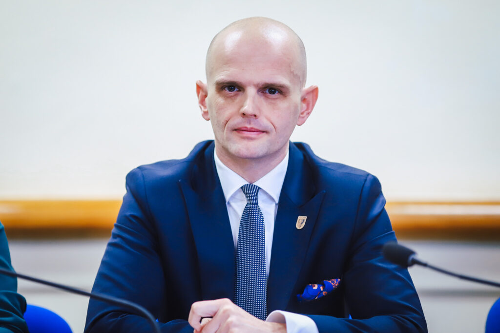 Mężczyzna w granatowym garniturze z krawatem w drobne białe kropeczki siedzi przy stole podczas XIX sesji Rady Powiatu Płockiego (kadencja 2024–2029), z widocznym mikrofonem na pierwszym planie.