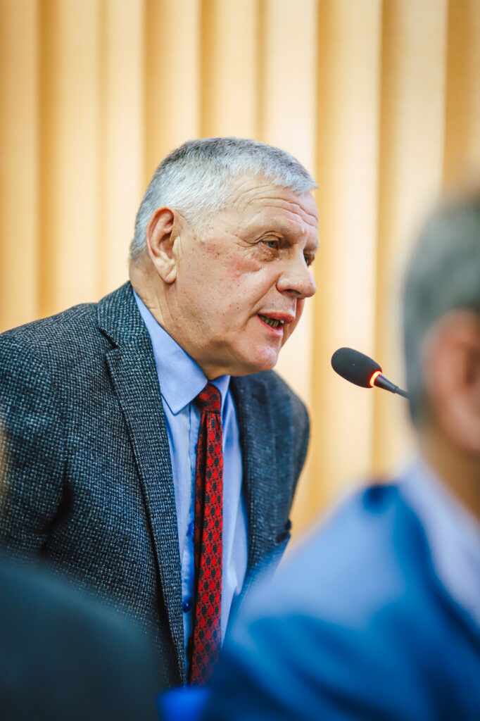 Uczestnik XIX sesji Rady Powiatu Płockiego przemawia do mikrofonu, stojąc przed żółtą zasłoną; mężczyzna w szarym żakiecie, błękitnej koszuli i czerwonym wzorzystym krawacie.