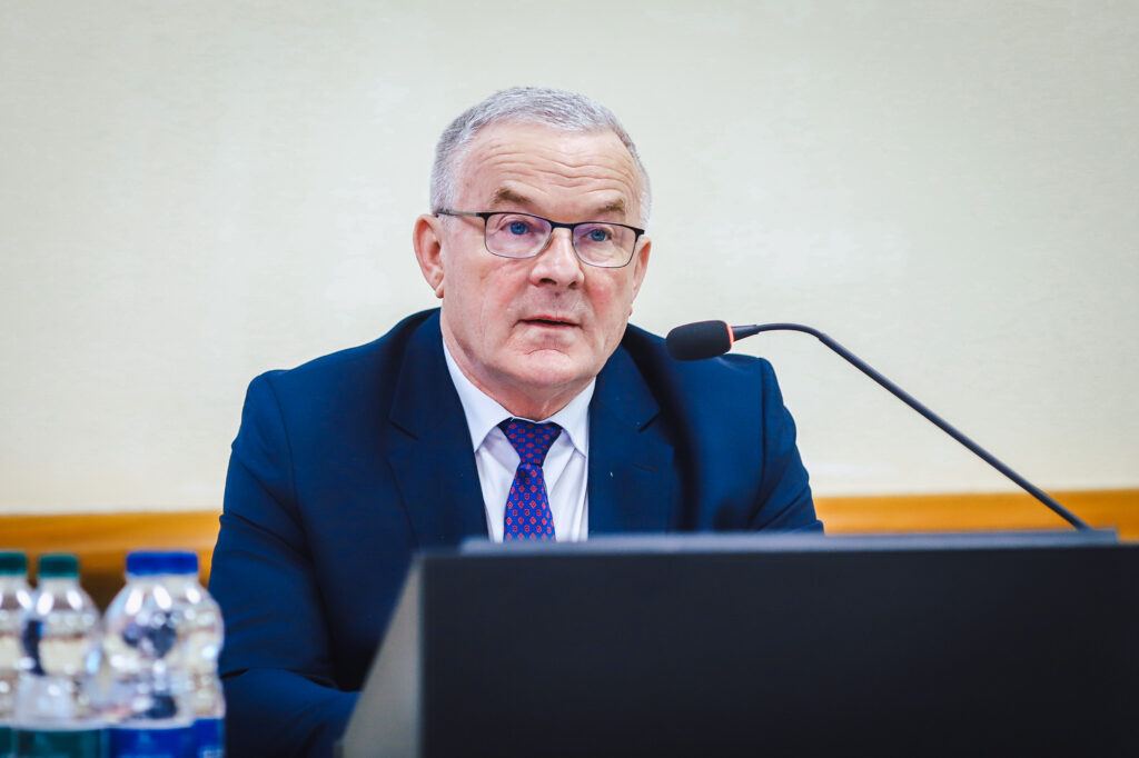 Mężczyzna w granatowym garniturze i okularach siedzi przy mikrofonie podczas XIX sesji Rady Powiatu Płockiego w kadencji 2024–2029.