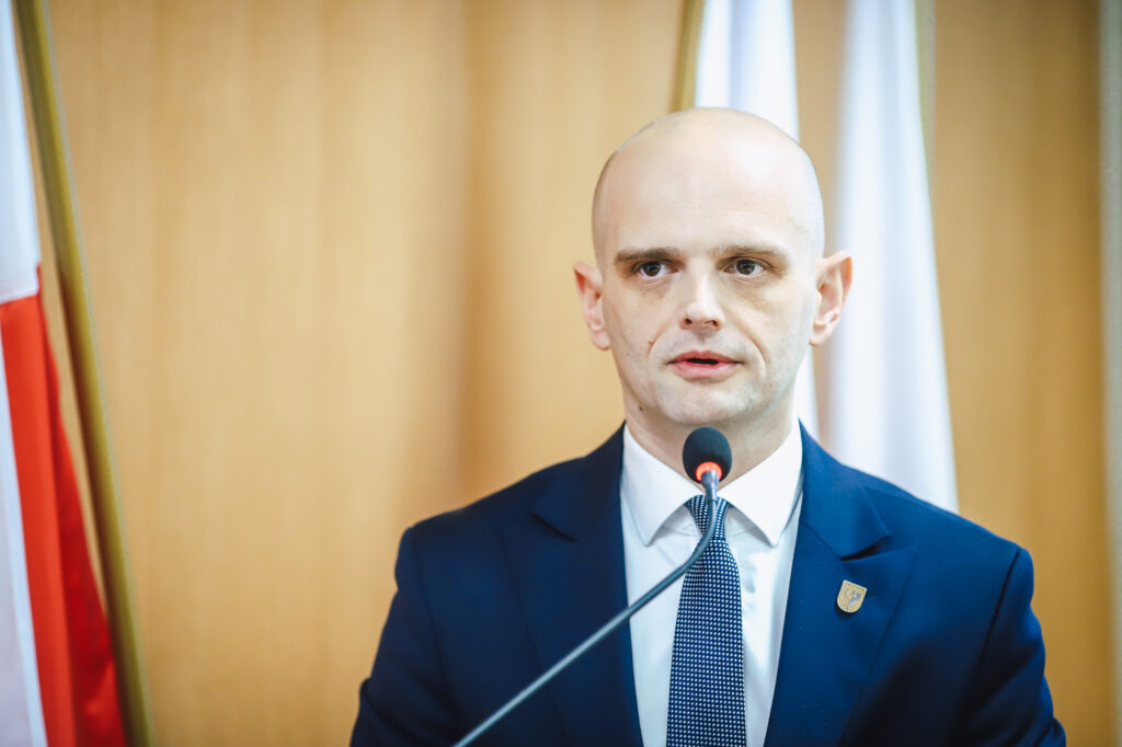 Mężczyzna w granatowym garniturze przemawia do mikrofonu podczas XIX sesji Rady Powiatu Płockiego.