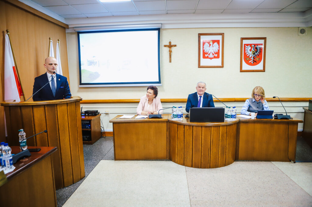 Mężczyzna w garniturze przemawia z mównicy podczas XIX sesji Rady Powiatu Płockiego w sali obrad; po prawej siedzą trzy osoby przy drewnianym stole z mikrofonami, a na ścianie widoczny krzyż i dwa herby państwowe.