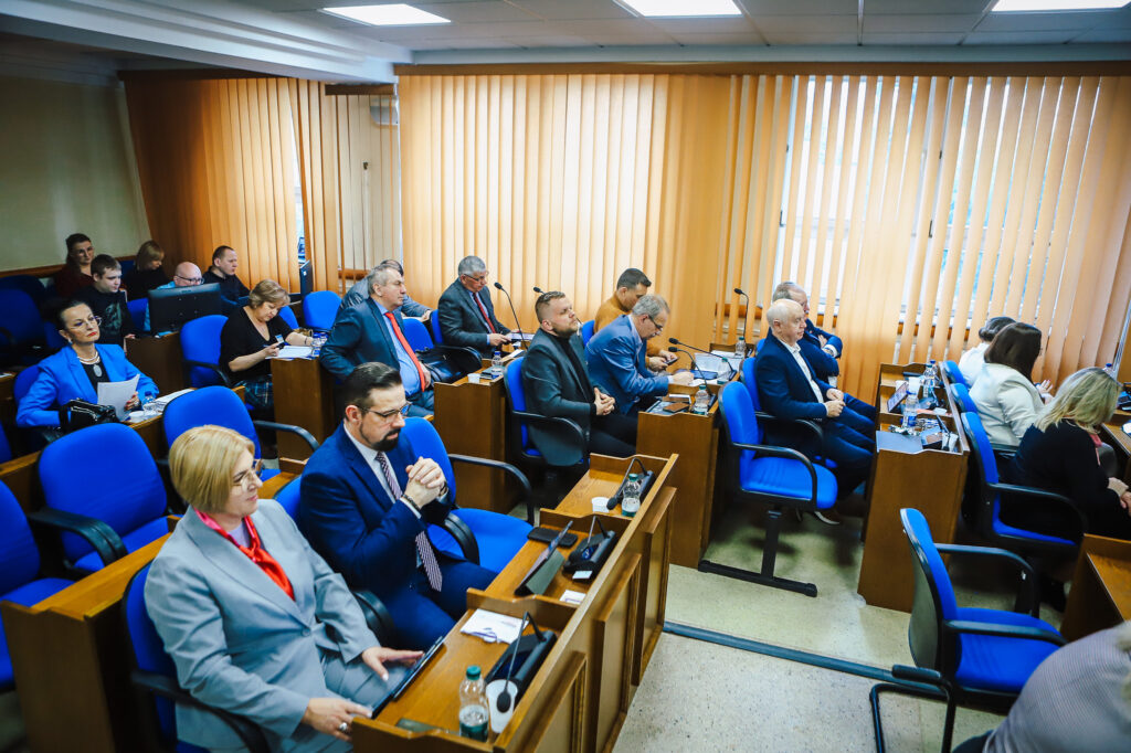 Uczestnicy XIX sesji Rady Powiatu Płockiego (kadencja 2024–2029) siedzą w sali obrad przy drewnianych pulpitach z mikrofonami i dokumentami, w tle żółto-beżowe rolety i niebieskie krzesła.