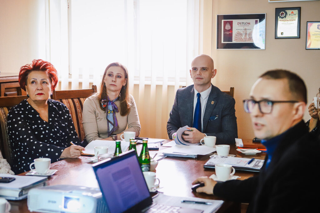 Grupa czterech osób siedzi przy stole w sali konferencyjnej, omawiając dokumenty podczas spotkania, z laptopem, filiżankami i butelkami wody na stole, a na ścianie widoczne są nagrody.