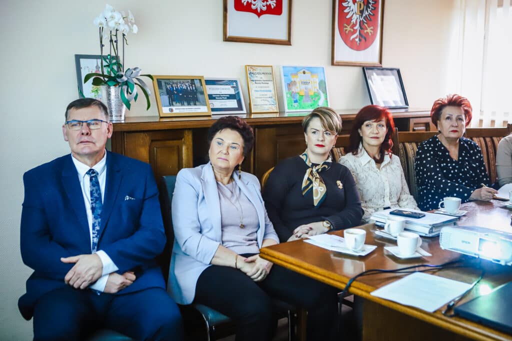 Grupa pięciu osób w formalnych strojach siedzi przy stole w sali konferencyjnej biura, omawiając wizualizacje nowej siedziby szkoły specjalnej w Goślicach.