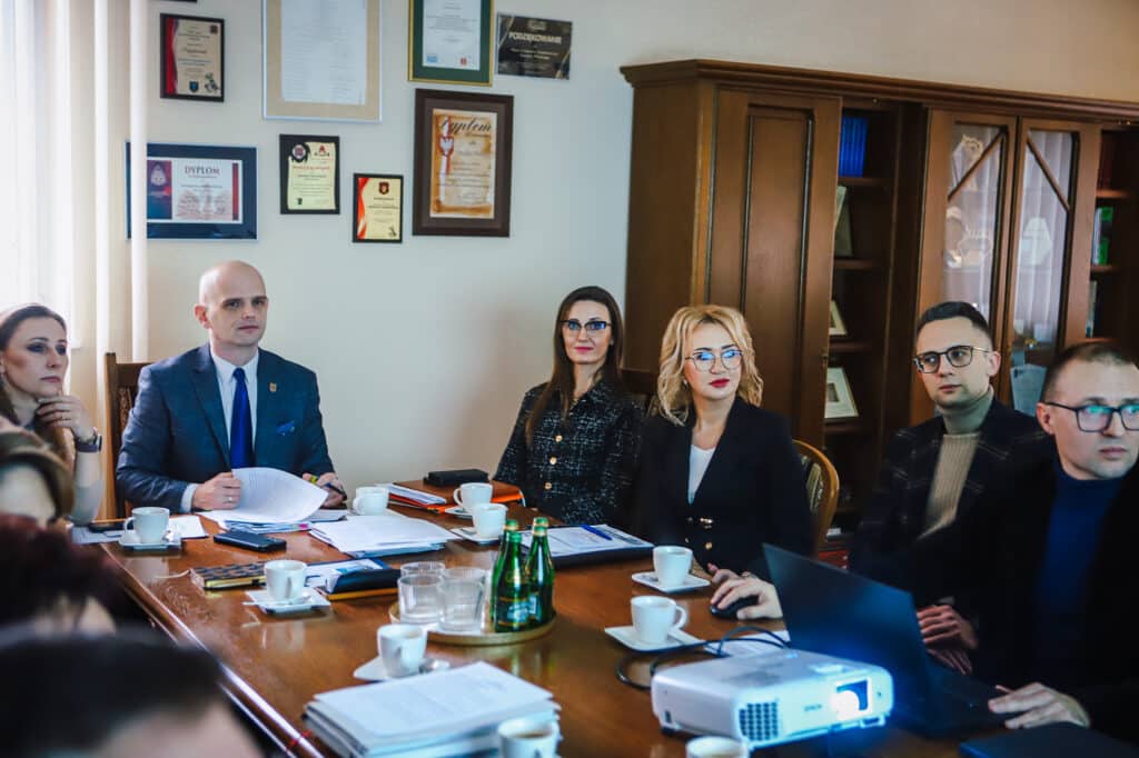 Grupa osób w formalnych ubraniach siedzi przy długim drewnianym stole w biurze, omawiając dokumenty i wizualizacje nowej siedziby szkoły specjalnej w Goślicach, przy projekcie i filiżankach na stole, w tle regał z dyplomami.