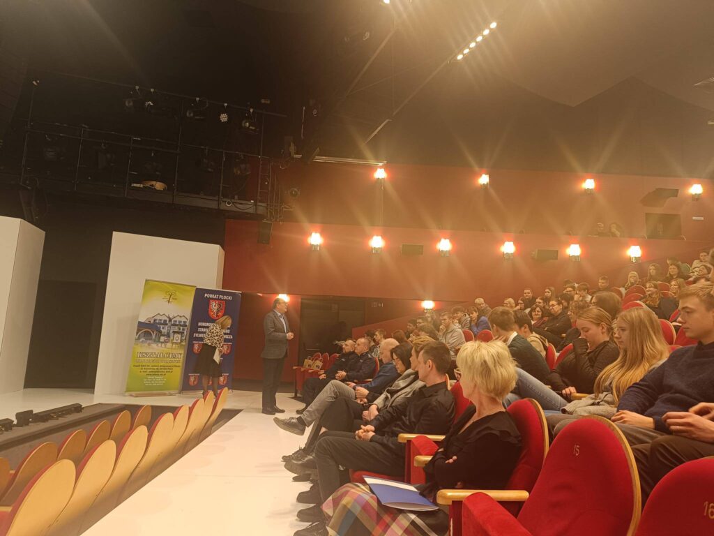 Mężczyzna w garniturze przemawia na scenie przed zgromadzoną publicznością podczas VI Powiatowego Dnia Profilaktyki „Świadomi dziś – bezpieczni jutro”.