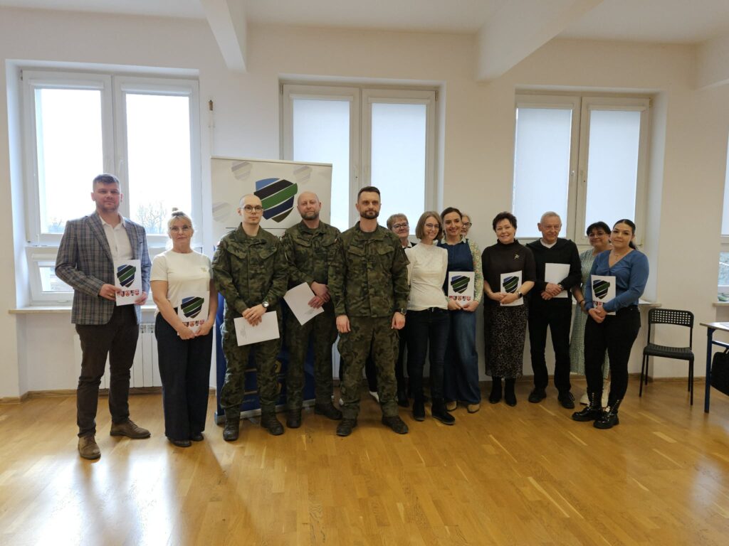 Grupa osób, w tym żołnierze w mundurach i cywile, stoi w jasnej sali po zakończeniu kwalifikacji wojskowej w Powiecie Płockim, wszyscy trzymają foldery z logo.