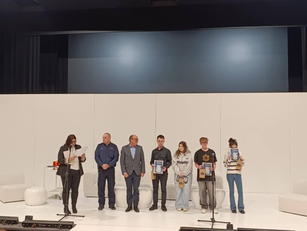 Na scenie podczas VI Powiatowego Dnia Profilaktyki „Świadomi dziś – bezpieczni jutro” stoi siedmioosobowa grupa: kobieta z mikrofonem i kartkami po lewej, a dalej mężczyźni i młodzi ludzie trzymają certyfikaty.