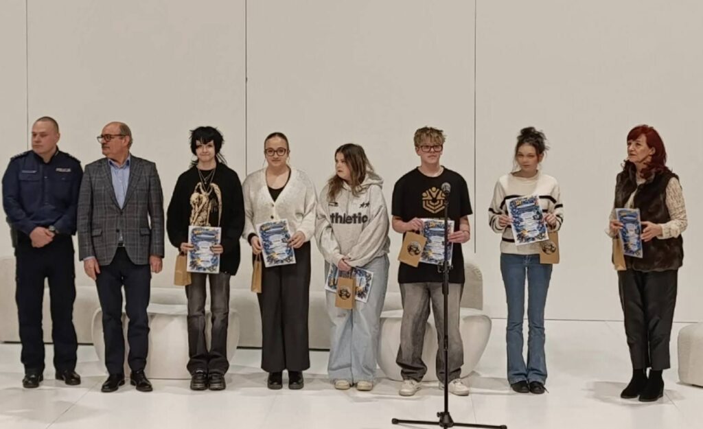 Grupa osób stoi na scenie podczas VI Powiatowego Dnia Profilaktyki „Świadomi dziś – bezpieczni jutro”, trzymając plakaty i torby z upominkami.