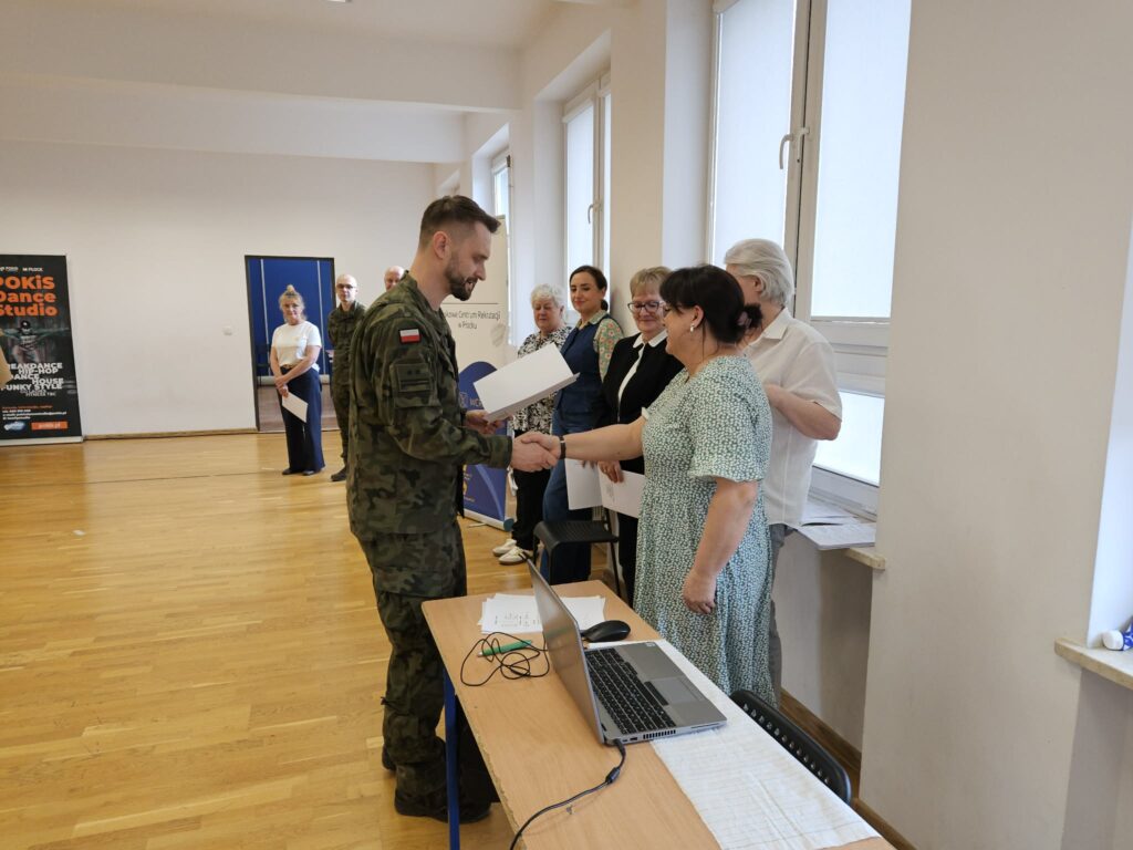 Żołnierz w mundurze wojskowym odbiera dokument od kobiety podczas zakończenia kwalifikacji wojskowej w Powiecie Płockim, w sali z laptopem na stole.