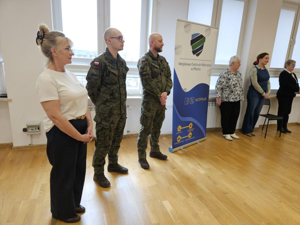 Grupa osób stojących w sali, w tym dwóch żołnierzy w mundurach, obok banera Wojskowego Centrum Rekrutacji w Płocku – zdjęcie z zakończenia kwalifikacji wojskowej w Powiecie Płockim.