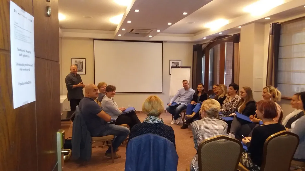 Grupa uczestników szkolenia dla przedstawicieli służb społecznych siedzi w sali konferencyjnej w półokręgu, a prowadzący stoi przy ekranie projekcyjnym.