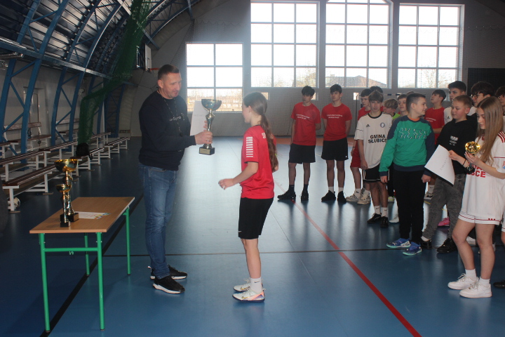 Mężczyzna wręcza puchar dziewczynie w czerwonej koszulce podczas ceremonii nagród na powiatowych finałach halowej piłki nożnej, w tle stoją inni młodzi zawodnicy.