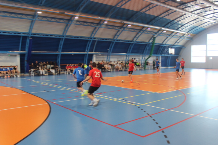 Podczas powiatowych finałów w halowej piłce nożnej zawodnicy w niebieskich i czerwonych strojach walczą o piłkę na boisku w hali, a w tle widać publiczność.