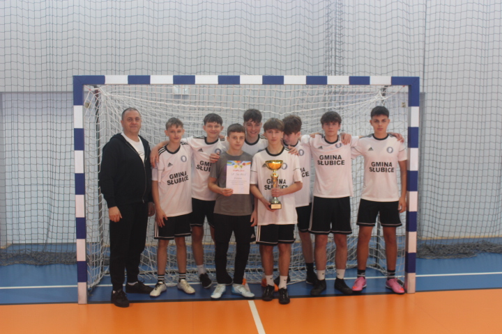Drużyna młodych piłkarzy w białych koszulkach z napisem Gmina Słubice stoi w hali sportowej przed bramką, trzymając dyplom i puchar po powiatowych finałach w halowej piłce nożnej.