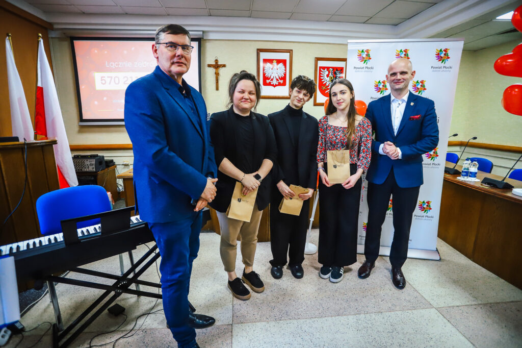 Pięć osób stoi na sali konferencyjnej podczas uroczystego podsumowania Finału WOŚP w powiecie płockim, na tle flag i banerów Powiat Płocki.