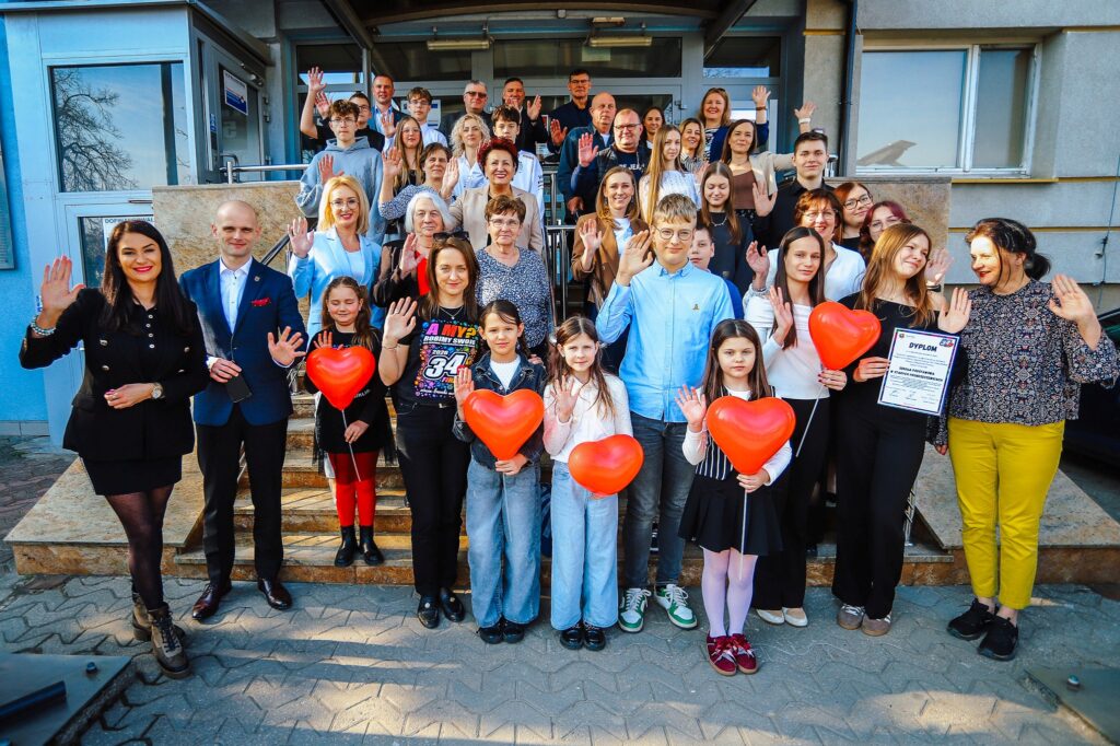 Grupa uczestników uroczystego podsumowania Finału WOŚP w powiecie płockim stoi na schodach budynku, machając do kamery, a część osób trzyma czerwone balony w kształcie serc.