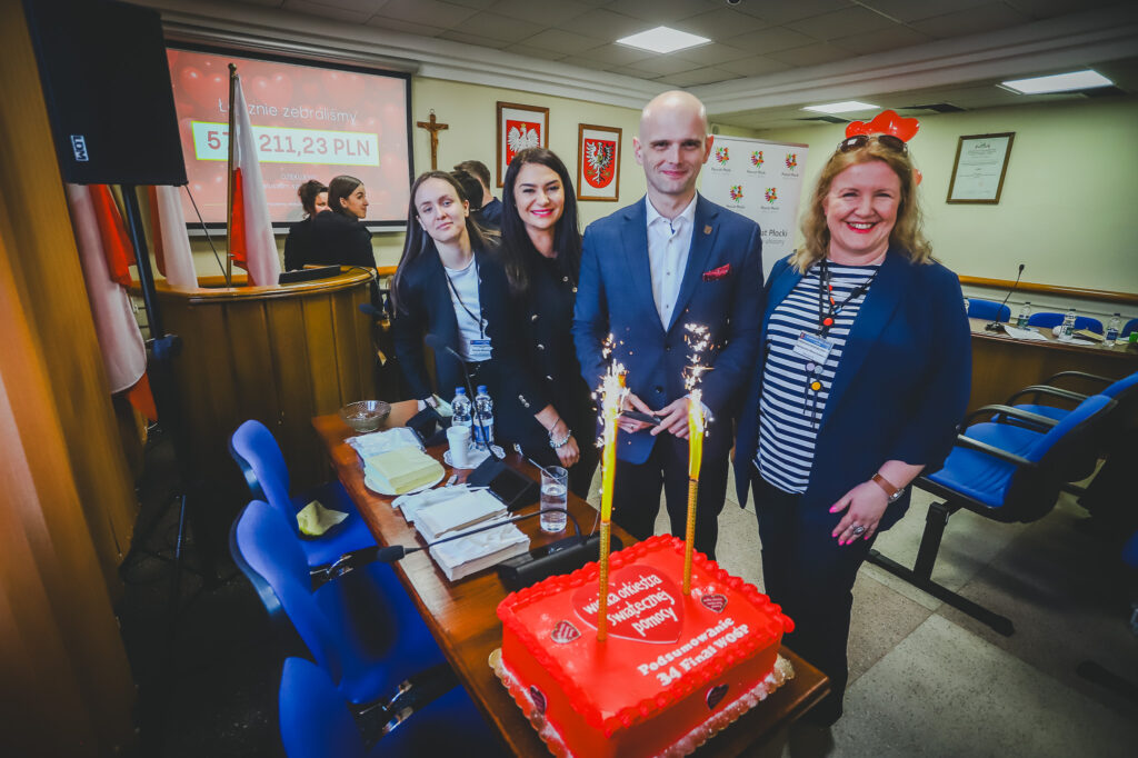 Grupa osób stoi przy stole z czerwonym tortem i iskryjącymi świecami, podczas uroczystego podsumowania tegorocznego Finału WOŚP w powiecie płockim.