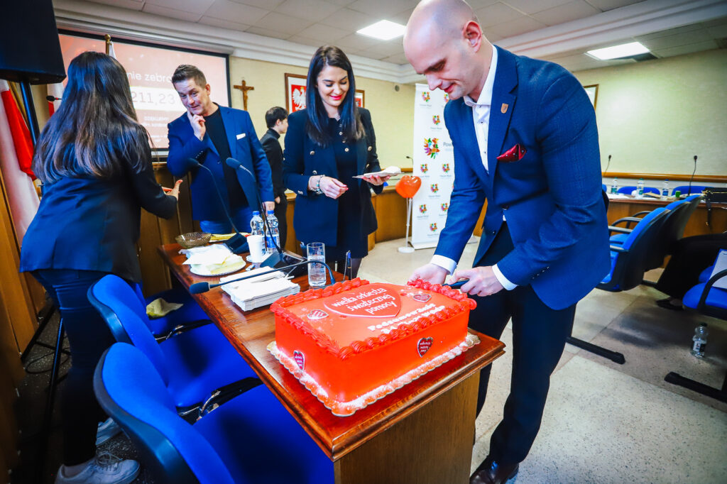 Uroczyste podsumowanie tegorocznego Finału WOŚP w powiecie płockim – grupa osób w formalnych ubraniach przy stole z dużym czerwonym tortem ozdobionym napisem WOŚP, podczas krojenia ciasta.