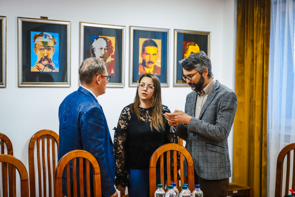 Trzy osoby stojące w sali z obrazami na ścianie prowadzą rozmowę podczas spotkania strażaków ochotników z przedstawicielami Fundacji ORLEN.