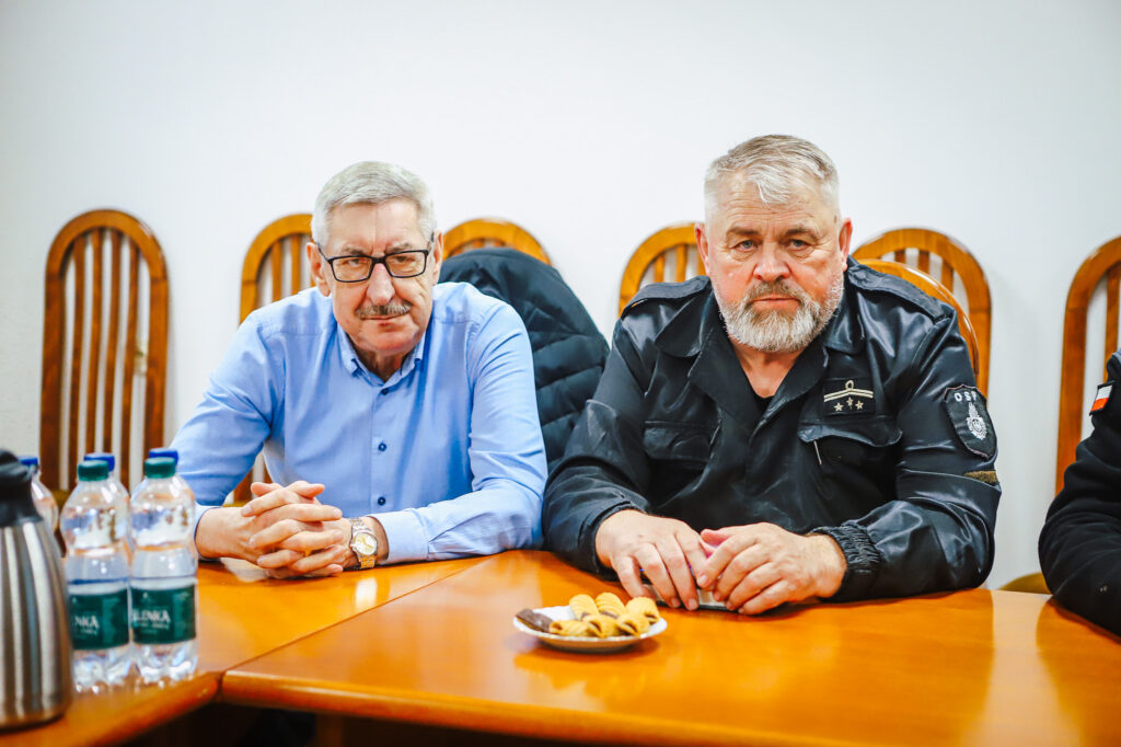 Dwóch mężczyzn, strażak ochotnik i przedstawiciel Fundacji ORLEN, siedzi przy stole podczas spotkania; na stole znajdują się butelki wody, metalowy termos i talerzyk ciastek.