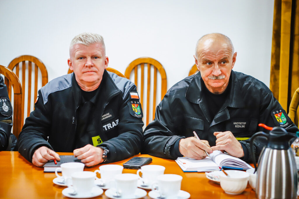 Dwóch strażaków ochotników w czarnych mundurach siedzi przy stole podczas spotkania z przedstawicielami Fundacji ORLEN.