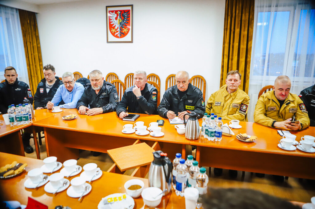 Grupa strażaków ochotników w mundurach siedzi przy długim stole w sali konferencyjnej podczas spotkania z przedstawicielami Fundacji ORLEN; na ścianie wisi herb.