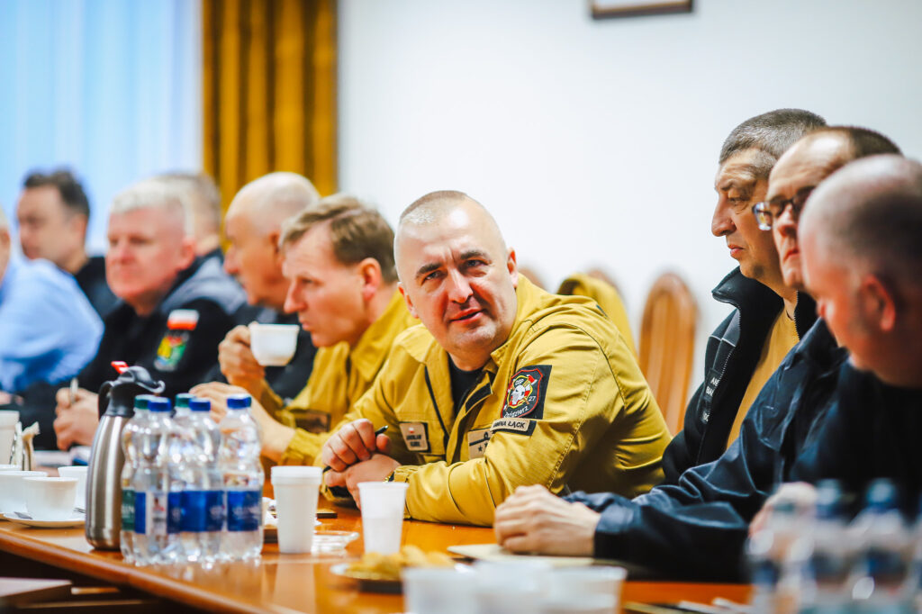 Grupa strażaków ochotników w mundurach siedzi przy stole podczas spotkania z przedstawicielami Fundacji ORLEN.