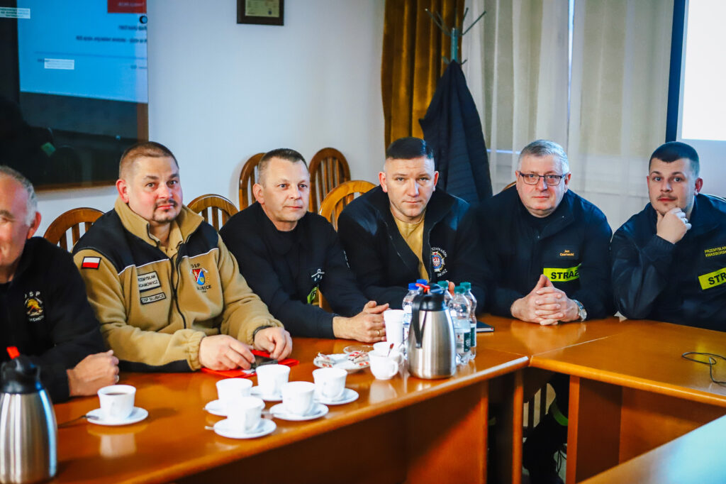 Grupa strażaków ochotników i przedstawicieli Fundacji ORLEN siedzi przy stole w sali konferencyjnej podczas spotkania, a na stole stoją filiżanki, kubki i butelki z wodą.
