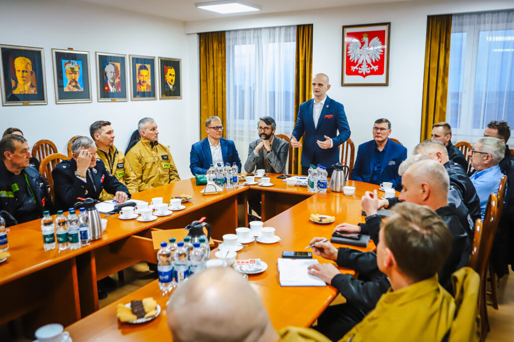 Grupa strażaków ochotników i przedstawicieli Fundacji ORLEN siedzi przy stołach ustawionych w literę U w sali konferencyjnej, gdy mężczyzna w granatowym garniturze wygłasza przemówienie.